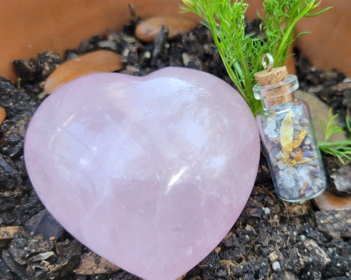 A rose quartz shaped like a heart sits in a potted plant next to a spell jar.
