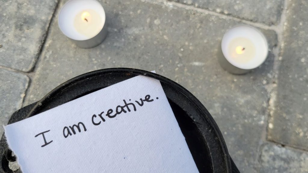 A piece of paper with the phrase, "I am creative" sits on top of a black cauldron with two lit candles in the background.