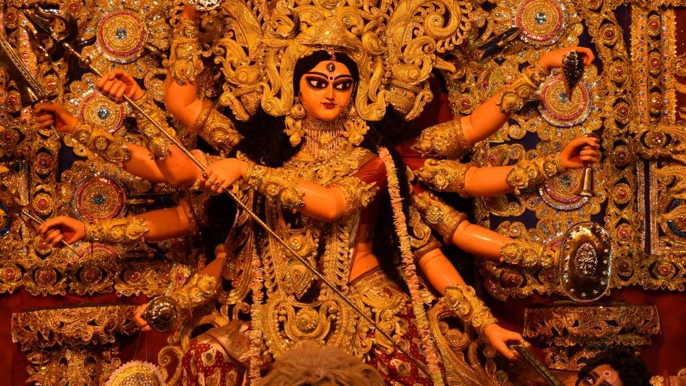 The Goddess Kali, a figure embellished with gold and bright colors. Kali looks to the side with 10 arms outstretched, each holding an object in her hand.